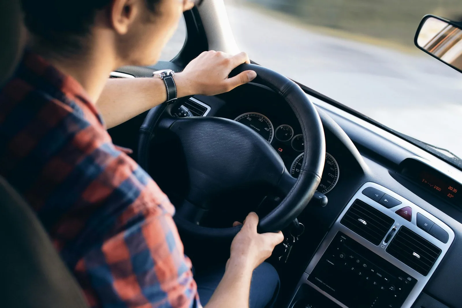 Auto Insurance Person driving a car with both hands on the steering wheel, wearing a plaid shirt.
